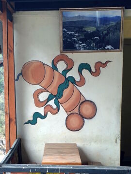 Zeichnung eines ejakulierenden Phallus' (samt zwei haariger Hoden) auf einer Hauswand, geschmückt mit einer grün-roten Stoffschleife rund um die Mitte des Penisschaftes